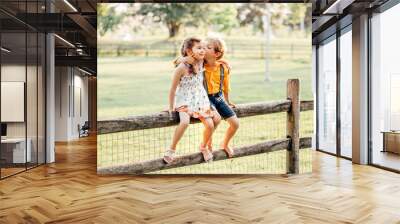 Two cute funny Caucasian children boy girl sitting on fence in park outside. Friends siblings kids hugging kissing at summer sunset. Romantic love and friendship natural lifestyle authentic moment. Wall mural