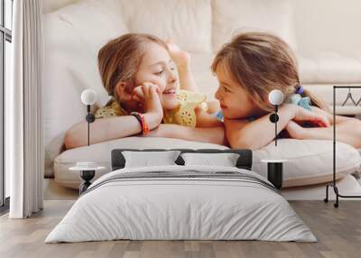 Two cute little Caucasian girls siblings playing at home. Adorable smiling children kids lying on a couch together. Authentic candid lifestyle domestic life moment. Happy friends sisters relationship. Wall mural