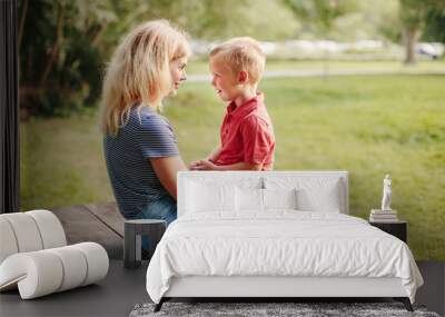 Young Caucasian mother and boy toddler son sitting together face to face. Family mom and child talking communicating outdoor on a summer day. Happy authentic family childhood lifestyle. Wall mural