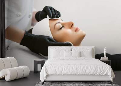 Close up of hands of skillful beautician cleaning and touching female face with cotton pad or sponge. The woman is lying and relaxing. Her eyes are closed with pleasure Wall mural