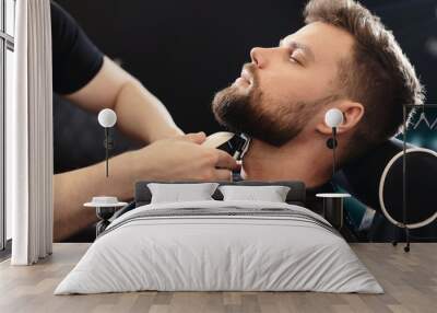 getting perfect shape. close-up side view of young bearded man getting beard haircut by hairdresser  Wall mural