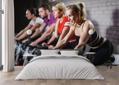 Group of smiling friends at gym exercising on stationary bike. Happy cheerful athletes training on exercise bike. Young men and woman working out at a class in the gym. Wall mural