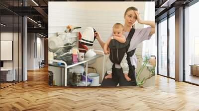 Tired multitasking mother and child in baby carrier. Young girl in the kitchen with a child. The difficulties of motherhood and loneliness Wall mural