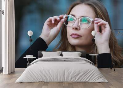 Myopia, close-up portrait of young woman student in eyeglasses for good vision looking up, blue building background Wall mural