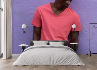 Smiling laughing African American man model posing in empty living coral color t-shirt standing against violet wall background Wall mural