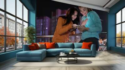 Young women friends looking at smartphone in the balcony terrace, night city background Wall mural