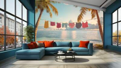 Vibrant swimsuits, bikinis, and lingerie hanging on a clothesline strung between two palm trees on a sandy beach, The sea and the evening sky before sunset on background Wall mural