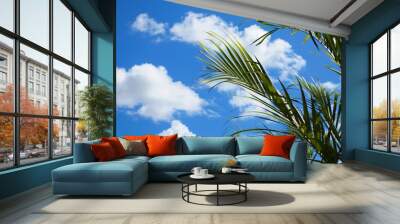 A green palm leaf against a bright blue sky with fluffy white clouds. Wall mural