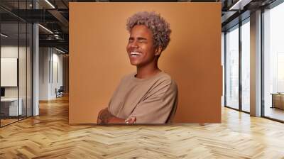 Overjoyed beautiful young African American woman smiles broadly and shows white teeth laughs happily, expresses sincere emotions looks aside wears beige t short, model against brown background. Wall mural