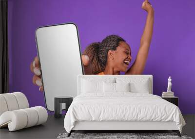 Young cheerful energetic African American woman screams with happiness holding phone with white screen and raises hand up experiencing euphoria after receiving SMS message stands on purple background. Wall mural