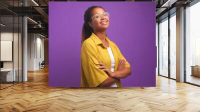 Young confident positive African American woman proudly posing with arms crossed in front of chest and raising chin looks at screen dressed in yellow shirt stands on purple studio background Wall mural