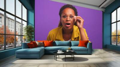 Young dazed African American woman grab glasses and open mouth in surprise when sees you or hears about huge discounts on shopping and sales in store stands on plain purple background Wall mural