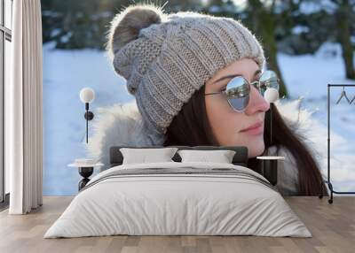 Portrait of a girl in a hat with a pompon against the background of a winter landscape. Beautiful woman 25 years old in sunglasses in a winter park. Wall mural