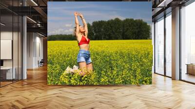 An adult happy woman in a red top and denim shorts jumps and makes a wheel element in a yellow rapeseed field. Motion blur effect. Wall mural