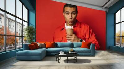 Busy young arabian man drink tea or coffee through straw. Look straight on camera and pose. Egyptian man hold paper cup in left hand. Isolated over red background. Wall mural