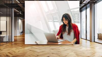 Calm and peaceful young woman working at table near window. Type on keyboard of laptop. Alone in room. Wear stylish red jacket. Productive day. Wall mural