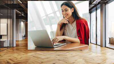 Cheerful attractive young woman working in cafe. Look on laptop and smile. Wear red jacket. Fashionable model typing on keyboard. Alone in room. Wall mural