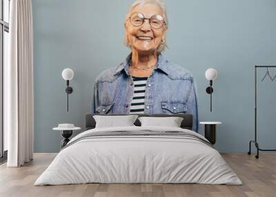 Cheerful very emotional old woman with gray hair wearing jeans jacket and shirt looking at camera, laughing and posing isolated over blue background. Smart happy senior lady. Happy feelings concept. Wall mural