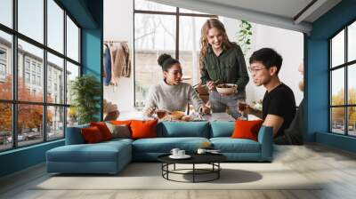 Cheerful woman holding bowl with salad in hand happily spending time with colleagues. Group of attractive international friends sitting at the table full of food having lunch together in cafe Wall mural