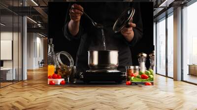 Cooking soup. A cook prepares soup in a saucepan against the background of ingredients. Delicious and healthy food. Black background. Salt, freezing in motion. Culinary and gastronomy. Wall mural