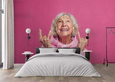 Emotional, cheerful and happy old woman in modern pink hoody posing on camera alone. Look up and laughing. Lady with grey hair show rock signs. Isolated over pink background. Wall mural