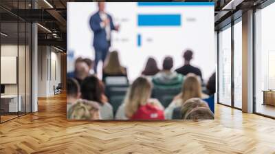 Audience attentively listening in a blurred seminar room, with presenter gesturing towards a data chart. Wall mural