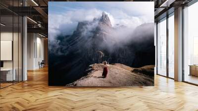 A beautiful girl in a red dress stands facing the mountain partially covered with clouds under the blue sky. Autumn. Italian Alps, Dolomites. Wall mural