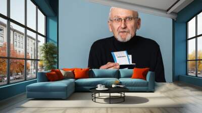 Old attractive man with gray hair and beard in eyeglasses and sweater holding tickets and passports in hands while amazedly looking in camera over blue background Wall mural