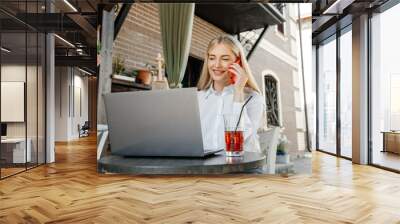 young beautiful blond caucasian woman sitting at table in outdoor terrace of cafe in summer day with notebook, businesswoman working, freelancer talking by phone and smiling Wall mural