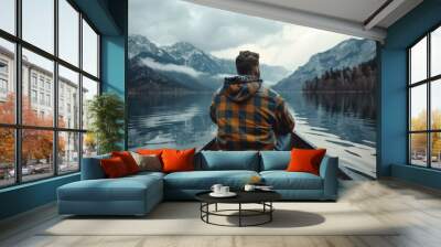 young man wearing a rugged flannel jacket and jeans sitting in a canoe on lake looking at distant mountains in background Wall mural