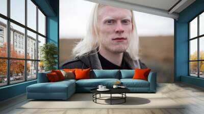 Portrait of a young, beautiful albino man with blue eyes and shoulder hair in a black quilted hooded coat, looking at the camera with a shrewd look against the background of the field. Wall mural