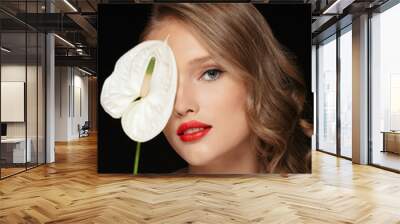 Portrait of young beautiful woman with wavy hair and red lips covering eye with white calla flower while dreamily looking in camera over black background Wall mural