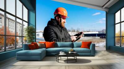 Pretty man - specialist civil engineer at orange hard hat is controls his project (outdoors). Blue sky and modern architecture (buildings) on background. Sunny day. Wall mural