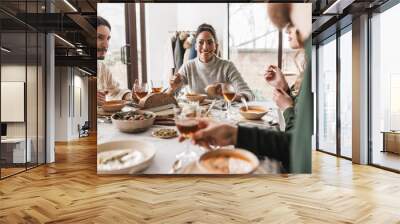 Smiling african american woman with dark curly hair sitting at the table eating soup with slice of bread in hand dreamily looking aside. Group of young international friends having lunch in cozy cafe Wall mural