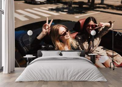 Two beautiful girls in sunglasses happily spending time together while driving cabriolet car on city streets outdoor Wall mural