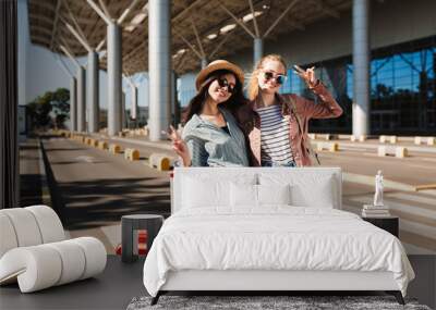 Two beautiful smiling girls in sunglasses happily looking in camera with red suitcase near airport Wall mural