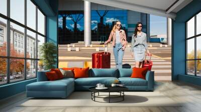 Two pretty girls in sunglasses happily walk along the pedestrian strip with red suitcases with airport on background Wall mural