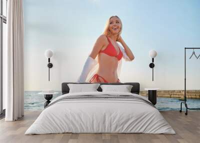 Young attractive blond woman in red swimsuit and white shirt happily standing alone on beach with sea on background Wall mural