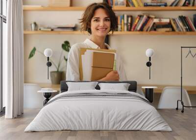 Young attractive smiling woman joyfully looking in camera with papers and notepad in office Wall mural