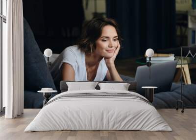 Young attractive smiling woman lying on sofa happily working on new project with laptop in office Wall mural