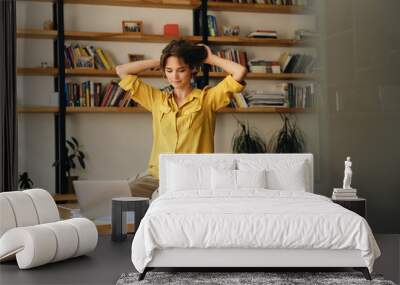 Young attractive woman in yellow shirt sitting on desk while dreamily working on laptop in modern office Wall mural