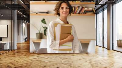 Young beautiful smiling woman dreamily looking in camera with papers and notepad in office Wall mural