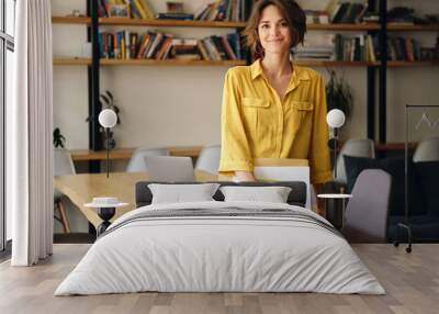 Young beautiful woman in yellow shirt leaning on desk with notepad and papers in hand while happily looking in camera in modern office Wall mural