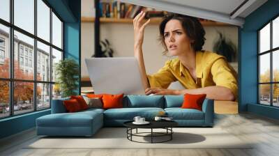 Young brunette woman indignantly sitting at table and working on laptop in office Wall mural