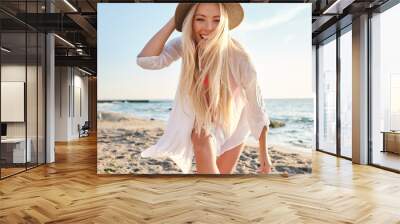 Young joyful blond woman in swimsuit and white shirt wearing hat while happily looking in camera with sea on background Wall mural