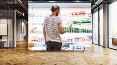 Young man in striped T-shirt thoughtfully choosing milk in dairy department of supermarket Wall mural