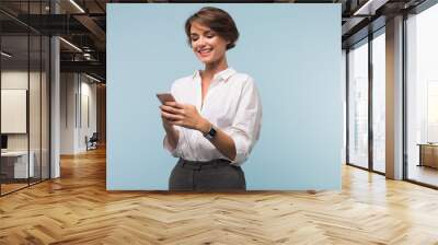 Young pretty smiling woman with dark short hair in white shirt happily using cellphone over blue background isolated Wall mural