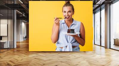 Young smiling woman in shirt happily looking in camera while eating chocolate cake over yellow background. Plus size model Wall mural