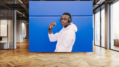 Young afro American black man wearing a white sweatshirt and headphones walking near a blue wall looking at camera. Modern look Wall mural