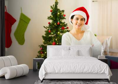 Cheerful woman in a santa hat smiling and posing with crossed arms in front of a festive christmas tree in her home Wall mural